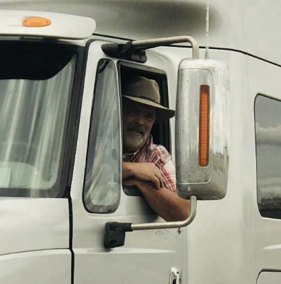 MikeMac leaning out the driver’s side window of a semi-truck, wearing a hat and smiling.